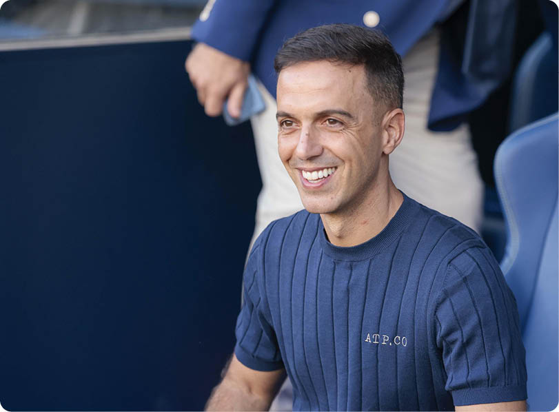 A person with short hair wearing a blue ribbed shirt is smiling while sitting in a sports stadium seat with a blurred person in the background. Descripci n generada con IA