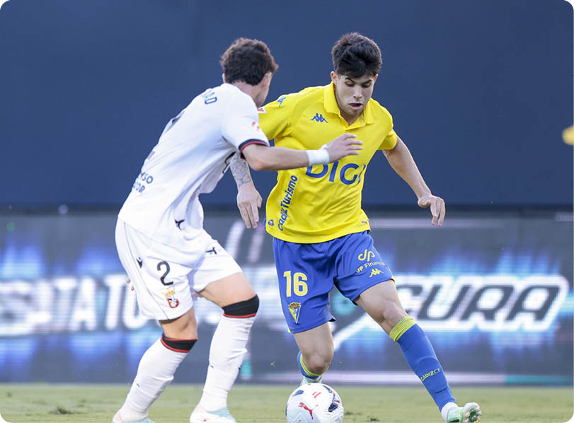 Two adults in sports uniforms are playing soccer on a grass field with one controlling the ball while the other defends. Descripci n generada con IA